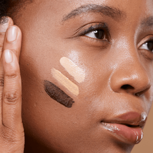 Jeune femme à la peau foncée et aux cheveux naturels bouclés appliquant des points de crème sur son visage, pot de soin vert en main. Image lifestyle authentique illustrant l'utilisation du kit soin pour peaux mélanines.